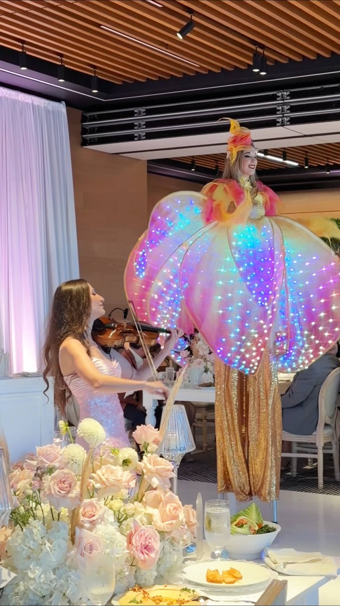 Tatev Muradyan playing violin beside a stilt performer in a glowing LED costume at an event