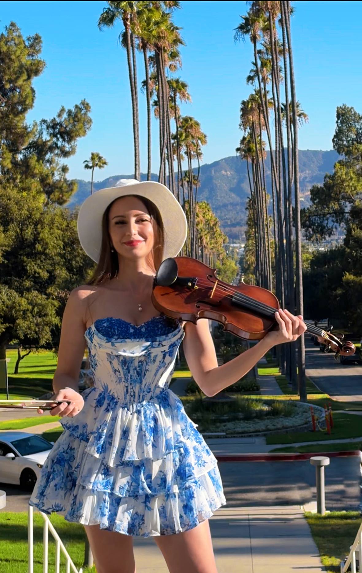 Tatev Muradyan in a blue floral dress playing violin on a palm-lined street