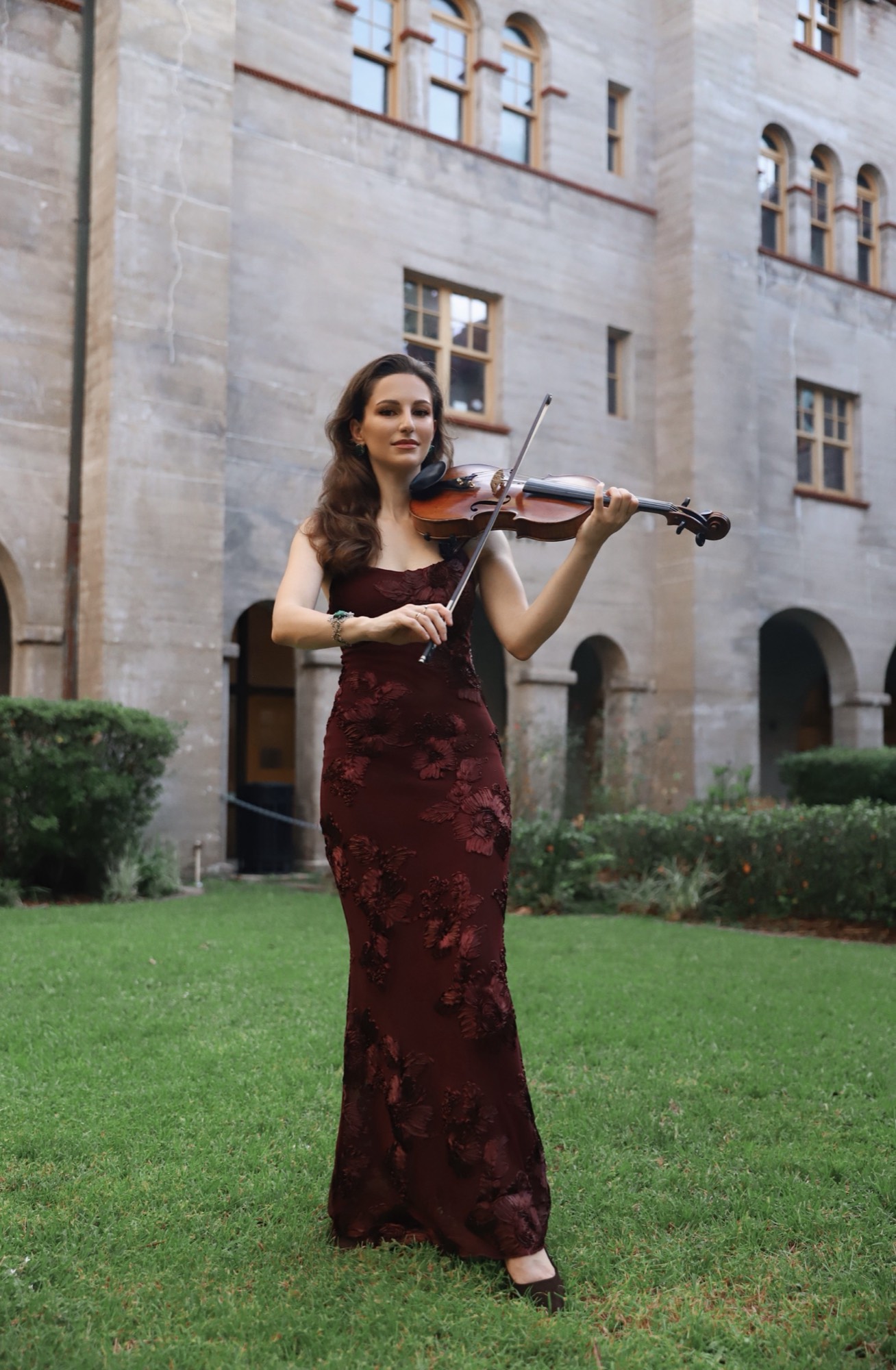 Tatev Muradyan in a burgundy gown playing violin on the lawn of a historic courtyard