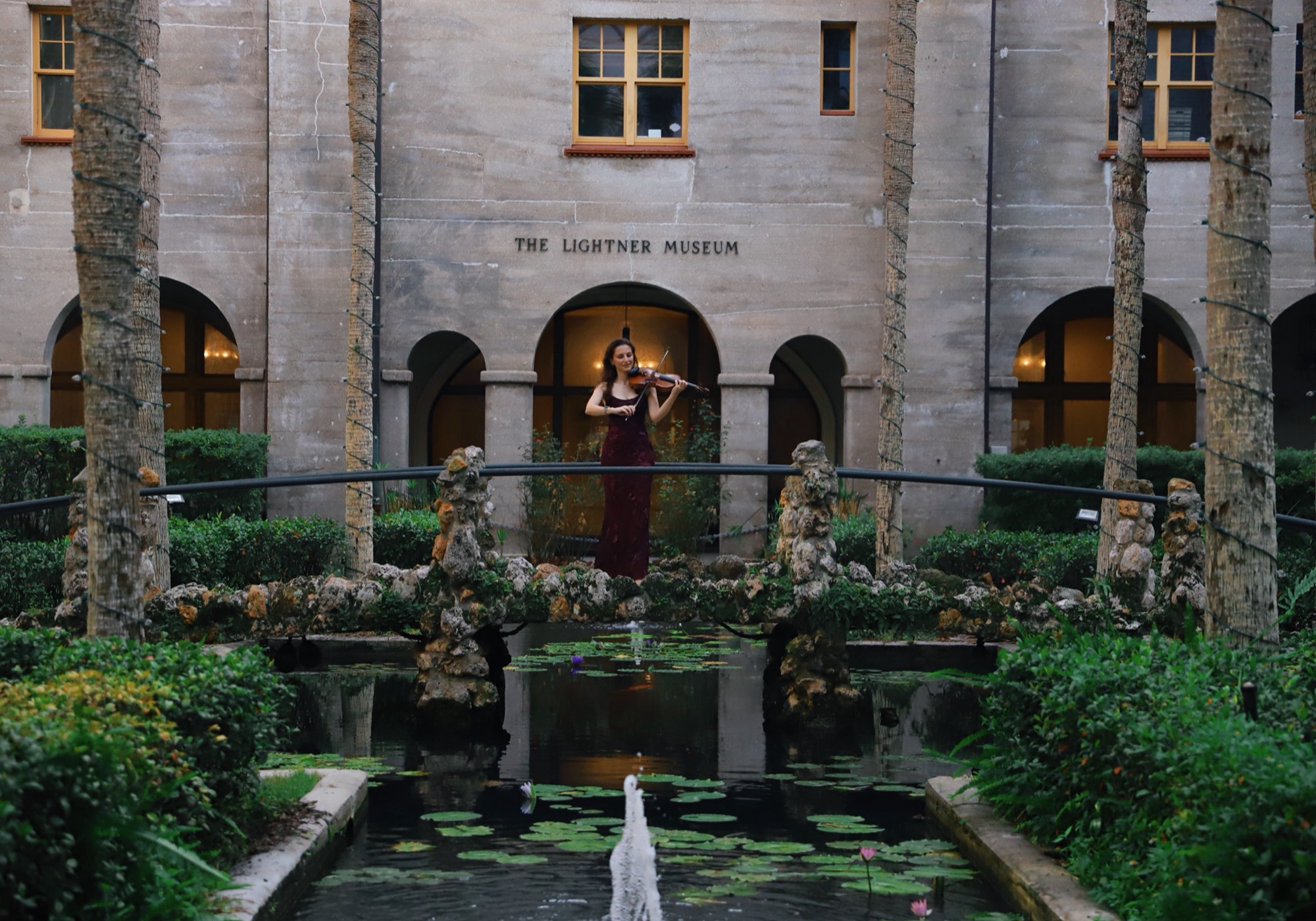 Tatev Muradyan playing violin on a stone bridge over a lily pond at the Lightner Museum