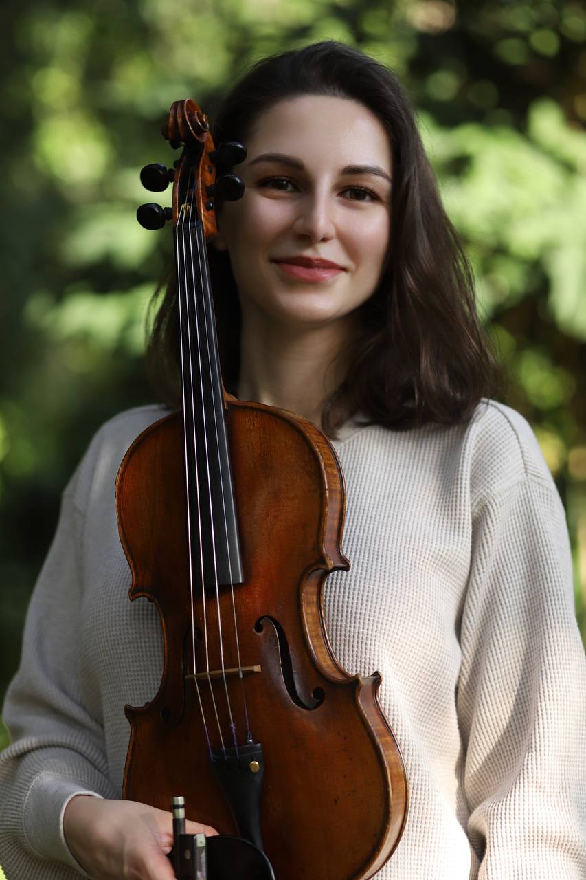 A close-up portrait of Tatev, a violinist with long, flowing brown hair and expressive dark eyes. She smiles warmly while looking directly at the camera, holding her violin gracefully in her hands. The background features a soft, blurred backdrop of lush green foliage from trees, creating a serene and natural atmosphere.