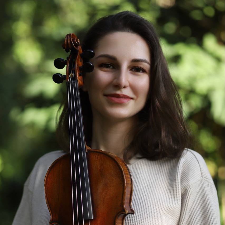 A close-up portrait of Tatev, a violinist with long, flowing brown hair and expressive dark eyes. She smiles warmly while looking directly at the camera, holding her violin gracefully in her hands. The background features a soft, blurred backdrop of lush green foliage from trees, creating a serene and natural atmosphere.